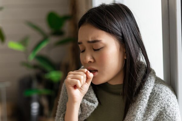 Sad ill asian girl staying on self quarantine during covid, catching flu and sitting at home with cup of tea near window, coughing in hand.