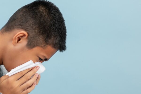 A boy is sneeze into tissue and feeling sick on blue background.